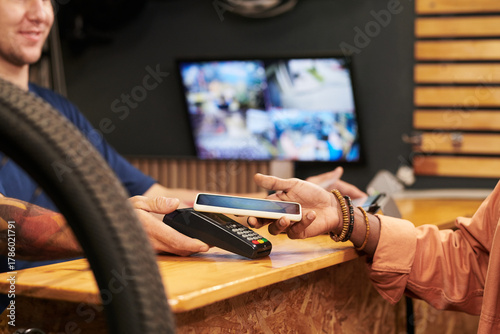 Young man holding smartphone near payment terminal while making contactless payment to young man at counter in retail store, security monitors visible in background