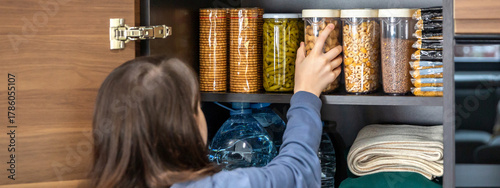 Unrecognizable girl storing peanuts container in emergency food storage pantry. 72 hour survival kit with water, cookies, pasta, beans, legumes, cereals and water ready for disasters or food shortage.