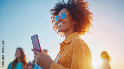 Vibrant young woman with afro hairstyle and stylish blue sunglasses, smiling joyfully while holding smartphone against warm, sun-kissed golden hour sky.