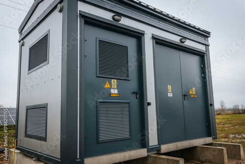 Modern electrical substation and power distribution unit installed on concrete base at a solar farm, with nearby transformers and solar panels visible in the background.