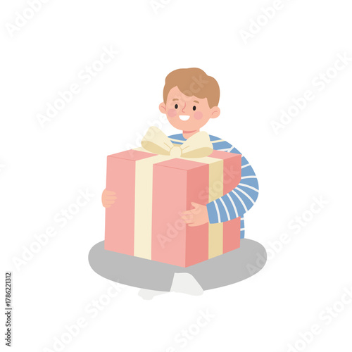 Young boy sitting cross legged holding a large pink wrapped gift box with a cream ribbon and bow wearing a blue striped shirt and gray pants with white socks