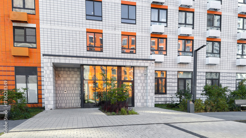 Entrance of modern residential building with geometric tiled facade in white and orange tones, large reflective windows, landscaped courtyard and urban design concept for real estate housing projects