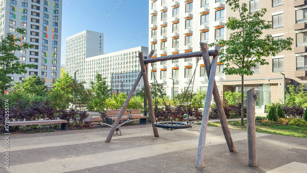 Fototapeta premium Urban playground with wooden swing and benches surrounded by modern residential buildings and greenery, symbolizing safe outdoor spaces and community lifestyle in contemporary city environments