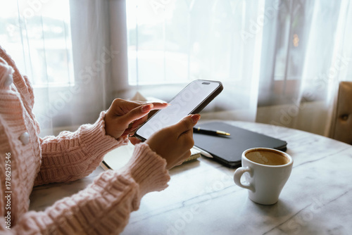 Woman using smartphone at cozy workspace with laptop, notebook, and coffee cup on marble table in soft daylight atmosphere.