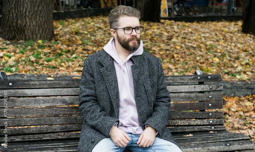 Fototapeta premium Man sitting on park bench in autumn leaves. Copy space and empty place for advertising. Calm atmosphere, reflection, and urban relaxation.