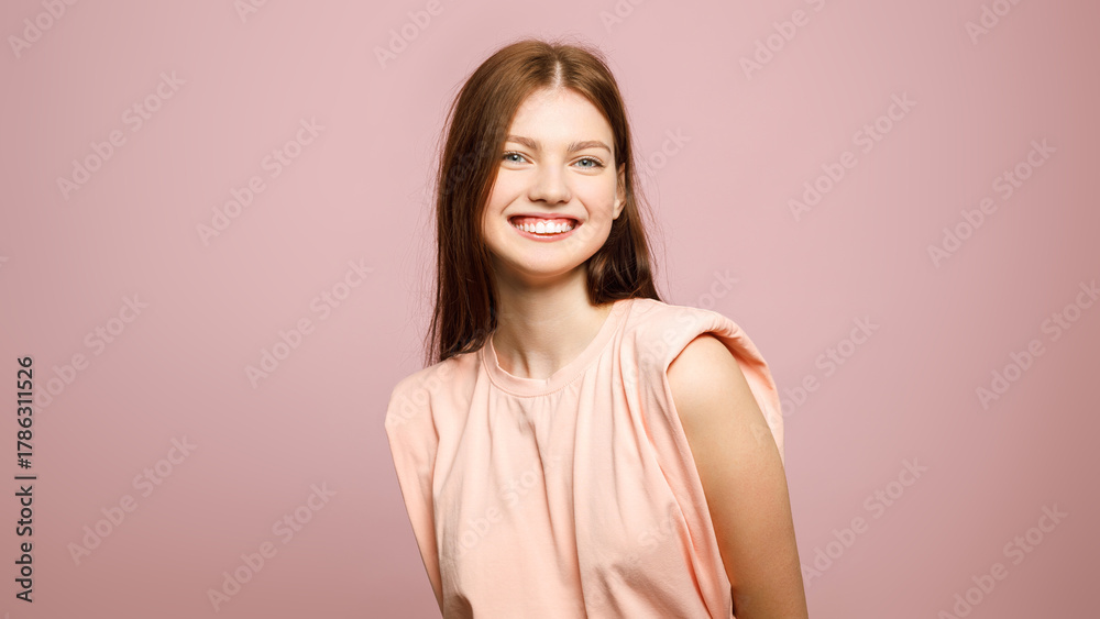 Obraz premium Cheerful young woman in a peach top smiling at camera against a pastel pink studio background with wide copy space.