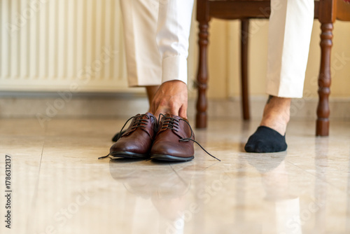 A man sits on a chair in a well-lit room, getting ready by placing on brown dress shoes while wearing one sock on his left foot. He appears focused and attentive.