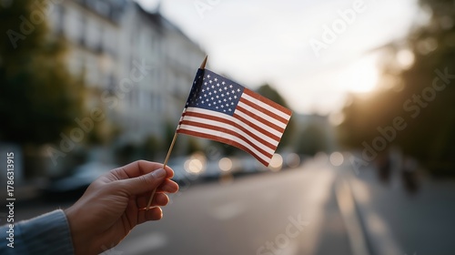 Fototapeta Naklejka Na Ścianę i Meble -  Hand holding a small USA flag while standing on a quiet morning street with soft sunrise light illuminating the surroundings — concept of patriotism, national pride, morning serenity, and lifestyle