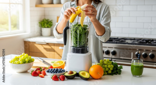 Fototapeta Naklejka Na Ścianę i Meble -  Woman preparing a fresh, healthy green smoothie with fruits and vegetables in a bright kitchen.