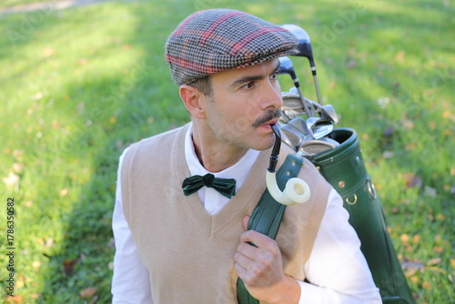 Фотография Elegantly dressed male caddie with mustache carrying golf bag across green course in classic vintage style