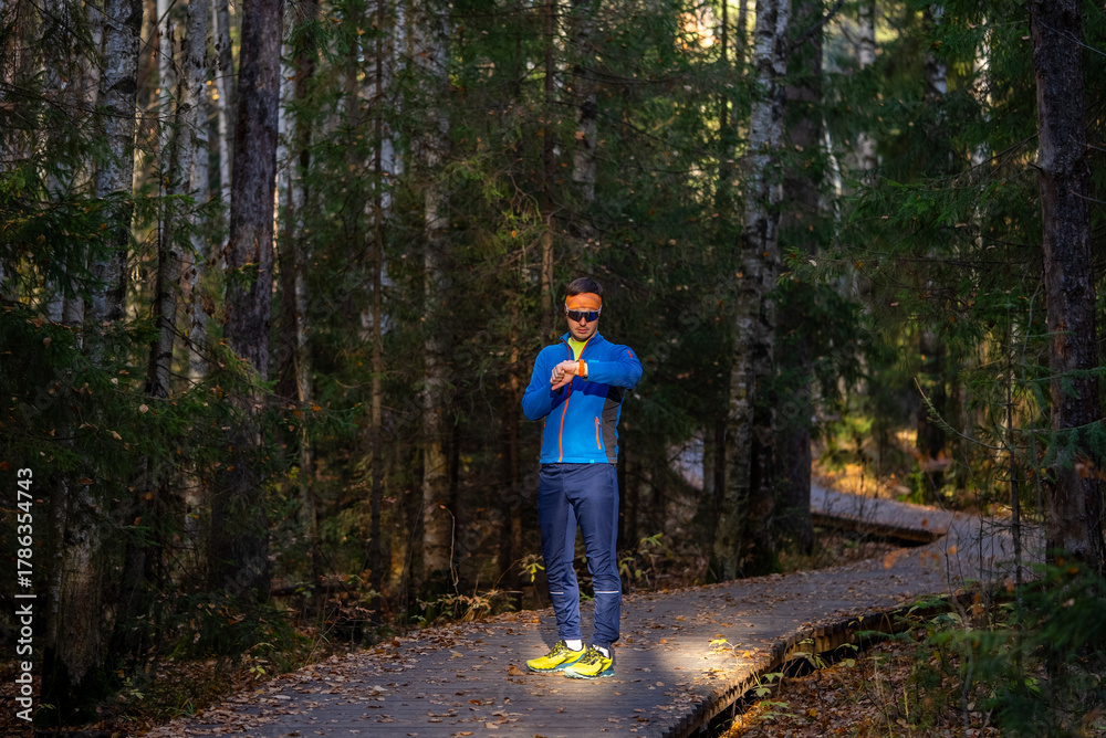 Fototapeta premium Man in athletic apparel is checking his smart watch after a training session on a wooden boardwalk trail surrounded by a dense forest, focusing on health and fitness data in nature