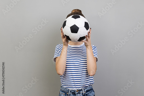 Woman hiding her face behind black and white soccer ball, watching football match.
