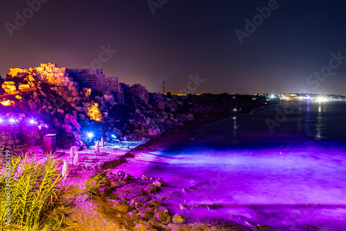 Fototapeta Naklejka Na Ścianę i Meble -  Magical night view of ancient fortress walls illuminated by blue and green lights on a beach, with the Mediterranean Sea and distant resort hotels. Side, Antalya, Turkey.

