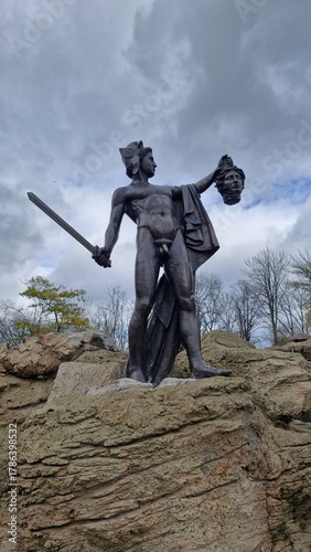 Autumn view of the “Perseus holding the head of Medusa” statue made of sandstone in Sofiyivka Park, Uman, Ukraine, with a cloudy gray sky in the background. Vertical mobile photo.