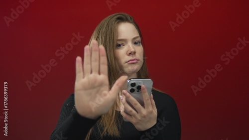 Woman raises palm and holds smartphone against red wall in studio with serious expression; rejection privacy.