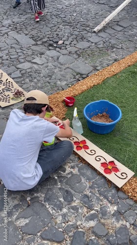 Person preparing colorful sawdust carpet on cobblestone street using stencil and natural materials during Holy Week celebration in Antigua, Guatemala.