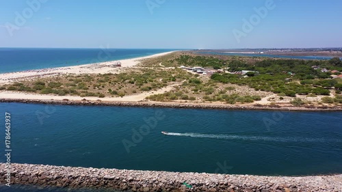 calm ria formosa channel with boat cruising along protected inlet captain steering wake past rocky breakwater marina and sand spit visible, clear blue water reflecting sky and distant Portugal Tavira