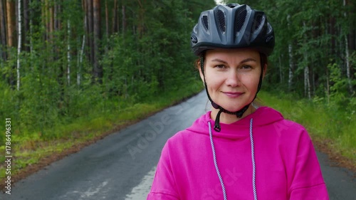 Portrait of a happy cyclist wearing a protective helmet and a pink hoodie, standing on an asphalt road in a beautiful green forest, enjoying her outdoor recreational activity and healthy lifestyle