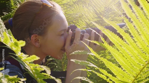 A young woman with a camera is surrounded by vibrant green ferns, capturing the natural beauty of her surroundings in a serene outdoor setting