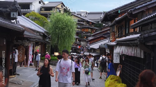 Tourists and locals move along stone paved Ninen zaka in Kyoto. Wooden machiya, lanterns, noren, and a willow line the hill. Wide shot with shallow depth of field.