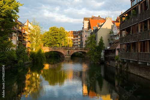 Golden hour over the Pegnitz in Nurnberg, Germany, glows warmly. The historic Maxbrücke spans the river like a calm anchor between leafy banks. Reflected facades amplify slow evening magic.