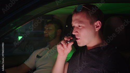 Two male friends sitting inside a car at night, one man vaping an electronic cigarette while the driver waits, creating a cloud of smoke with a modern alternative to traditional tobacco