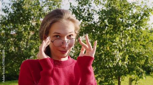 Playful young woman wearing a red sweater and round eyeglasses making funny faces and looking at the camera while adjusting her glasses in a park on a sunny day with green trees behind her.