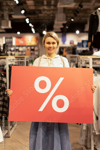 The girl is holding a big red banner with a percent sign and smiling. The girl in the clothing store shows a big discount, sale. Black Friday