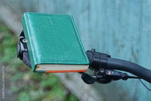Fototapeta Naklejka Na Ścianę i Meble -  one small closed green notepad with leather cover lies on a black bicycle handlebar on a summer street