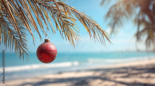 Fototapeta Naklejka Na Ścianę i Meble -  Single red Christmas ornament hanging from palm tree branch. Tropical beach panorama background
