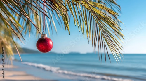 Fototapeta Naklejka Na Ścianę i Meble -  Single red Christmas ornament hanging from palm tree branch. Tropical beach panorama background
