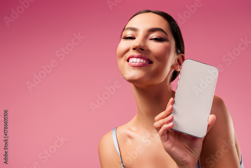 Gorgeous young woman holding smartphone in studio against pink background showcasing style and modern technology
