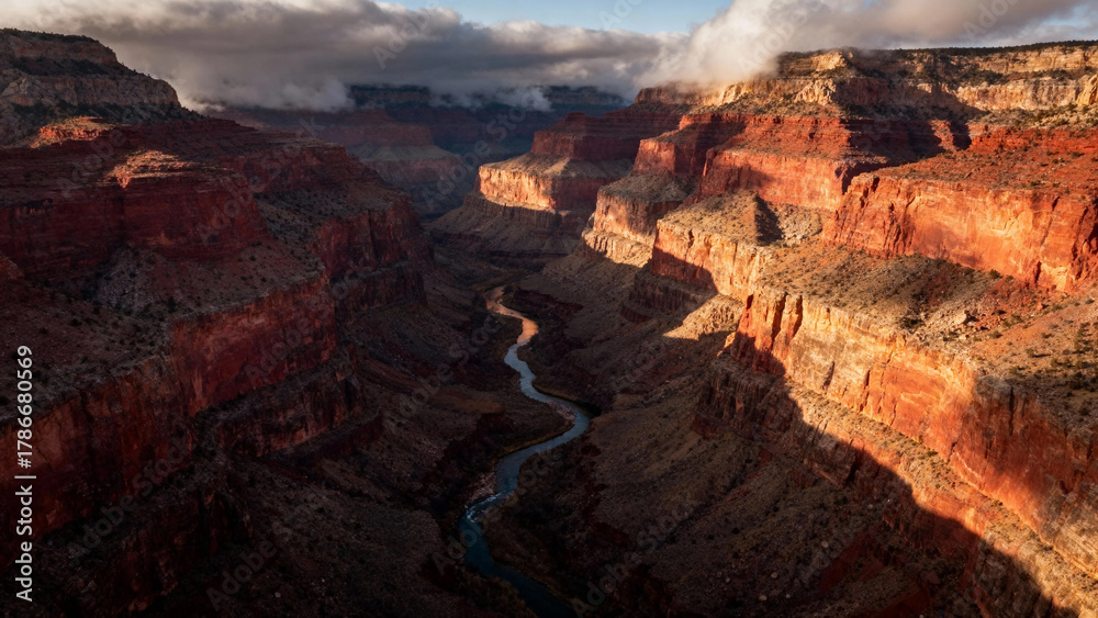 Obraz premium Aerial view of a deep canyon with layered rock formations and a winding river below, illuminated by sunlight under a partly cloudy sky.
