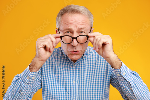A mature man is squinting and looking directly at the camera while adjusting his eyeglasses. The vibrant yellow wall behind him highlights his ophthalmic challenges.