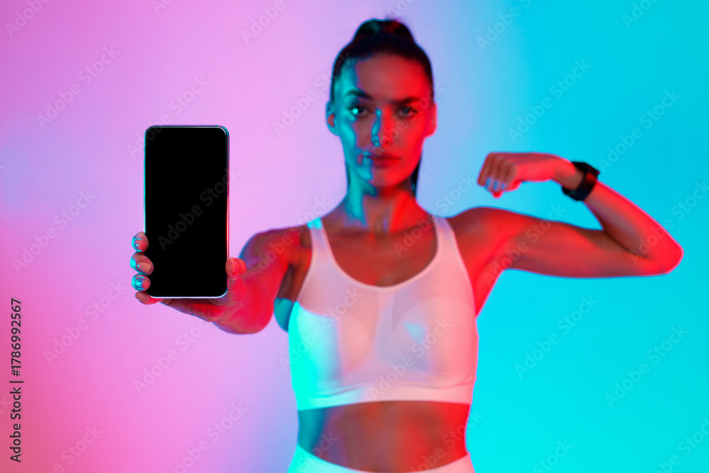 Obraz premium A fit woman stands in a colorful space with bright blue and pink lights. She holds a smartphone towards the camera and flexes her arm to display her strength.