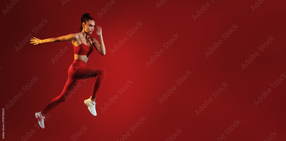 Naklejka premium A young woman in athletic wear leaps forward with determination against a solid red backdrop. Her focus and movement highlight the energy and strength of her workout.