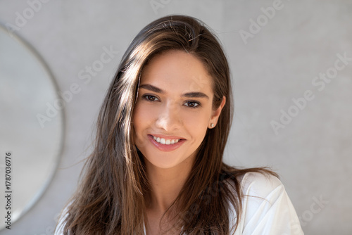 A young lady with smooth skin and thick hair smiles while looking at the camera in a bright bathroom at home. She radiates confidence and happiness, appreciating her natural beauty.