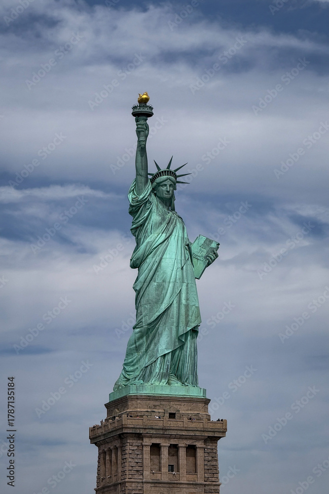 Fototapeta premium Iconic green monument of a woman holding a torch, standing on a stone pedestal against a cloudy sky in New York City.
