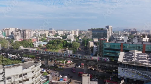 Aerial 4K shot of Mumbai Metro train entering Marol Naka station with cityscape and suburban buildings in view on a bright sunny day, showing modern urban infrastructure.