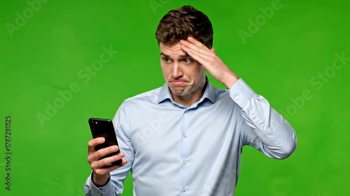 Man with confused expression looking at his phone against a green background.