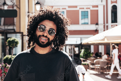 Handsome smiling hipster  model. Sexy unshaven Arabian man dressed in summer clothes. Fashion male with long curly hairstyle posing in the street at sunset. Cheerful and happy. In sunglasses