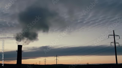 Silhouette of industrial smokestack and powerlines against a sunset sky with layered clouds and distant rolling hills