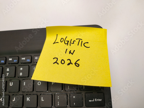 The image shows a yellow sticky note on a black computer keyboard. The note has the handwritten text LOGISTIC IN 2026
