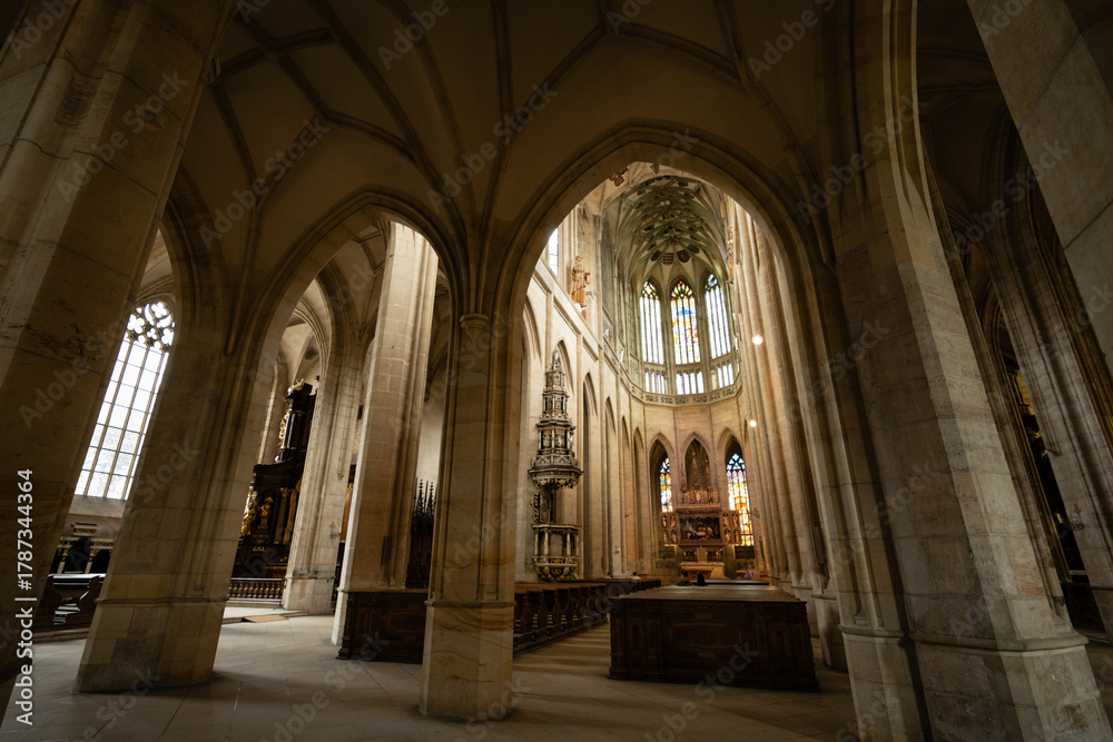 Fototapeta premium Kutná Hora, Czech Republic – August 8, 2025: Side aisle view of St. Barbara’s Cathedral with Gothic arches framing the ornate Baroque altar and chancel in Kutná Hora