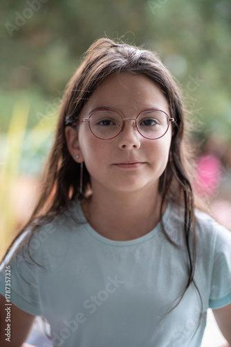 Vertical portrait of a girl in glasses wearing a light blue T-shirt, blurred natural background, soft daylight, copy space. Concept of innocence, calmness