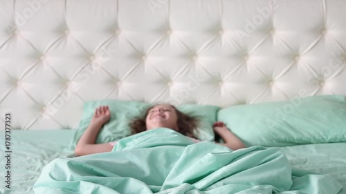 A little girl sits on a bed in pajamas and smiles at the camera, and than falls on the bed in slow motion