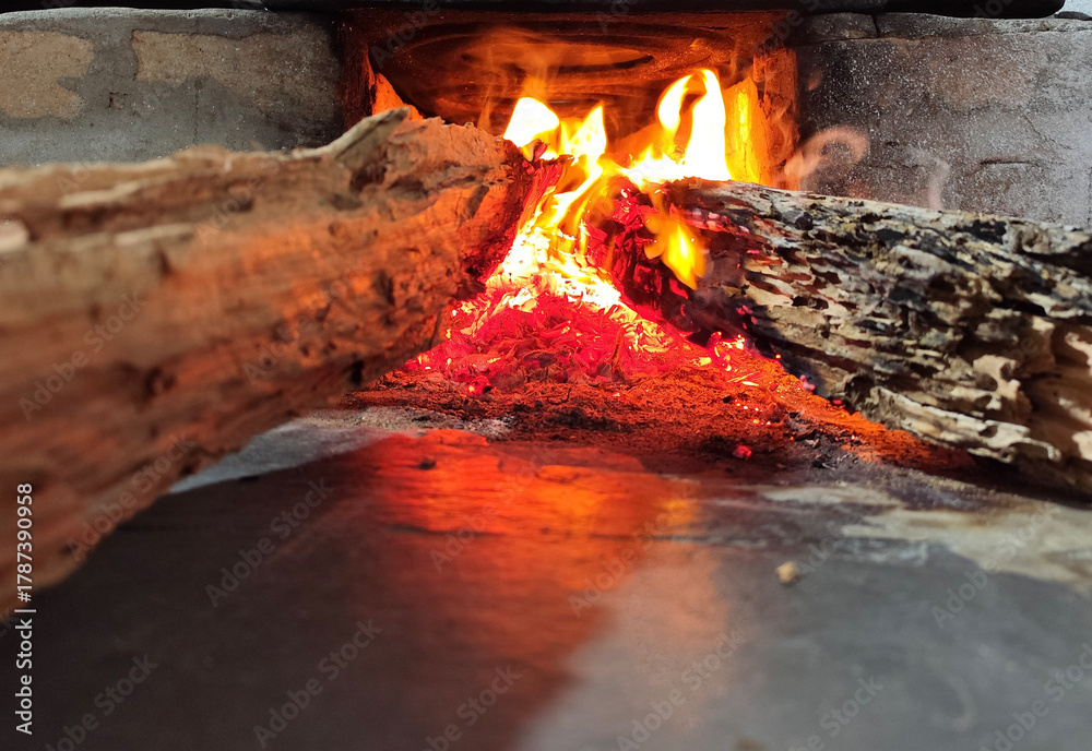 Naklejka premium Burning firewood inside a rustic fireplace with warm orange flames and glowing embers. Perfect for concepts of warmth, comfort, coziness, rustic lifestyle, and winter evenings.