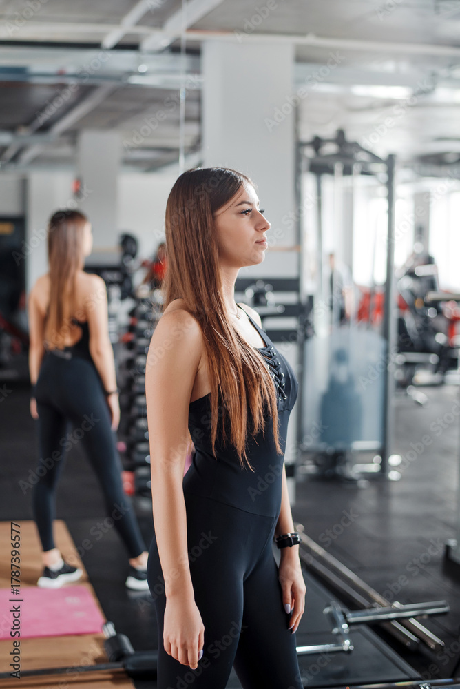 Fototapeta premium Healthy and fit young woman dressed in black sport clothes posing at modern gym. Caucasian female with long brown hair spending leisure time for training indoors.