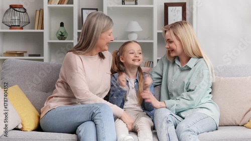 A senior woman grandmother, a young mother and a small granddaughter are sitting on the sofa, hugging, happy to meet. Three generations together, family coziness, Mother's Day celebration.