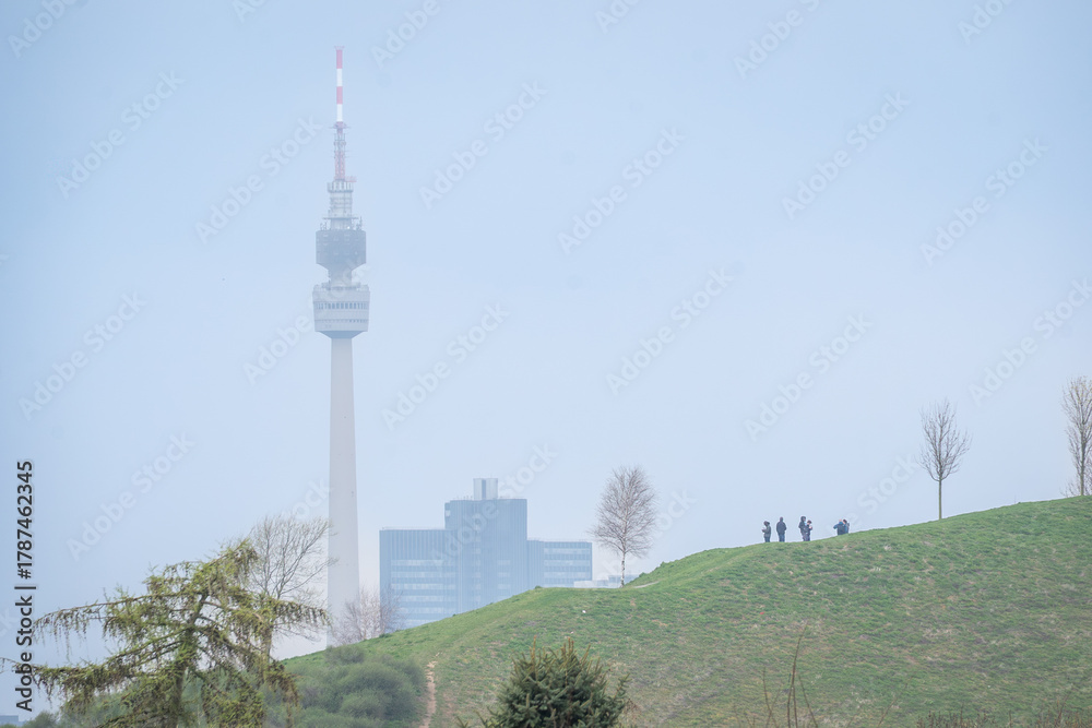 Obraz premium Urban Dortmund landscape with communication tower and silhouettes of people on grassy hill under soft blue sky.